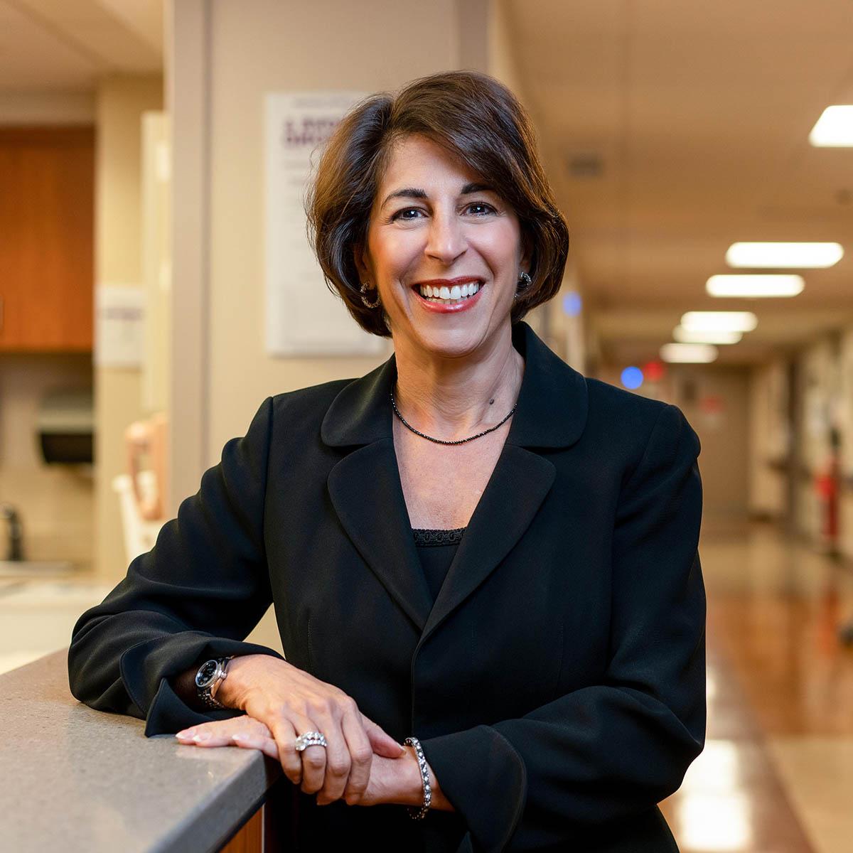 Photo of Sandra Rader, a smiling white woman with short brown hair, leans against a desk in a hospital corridor